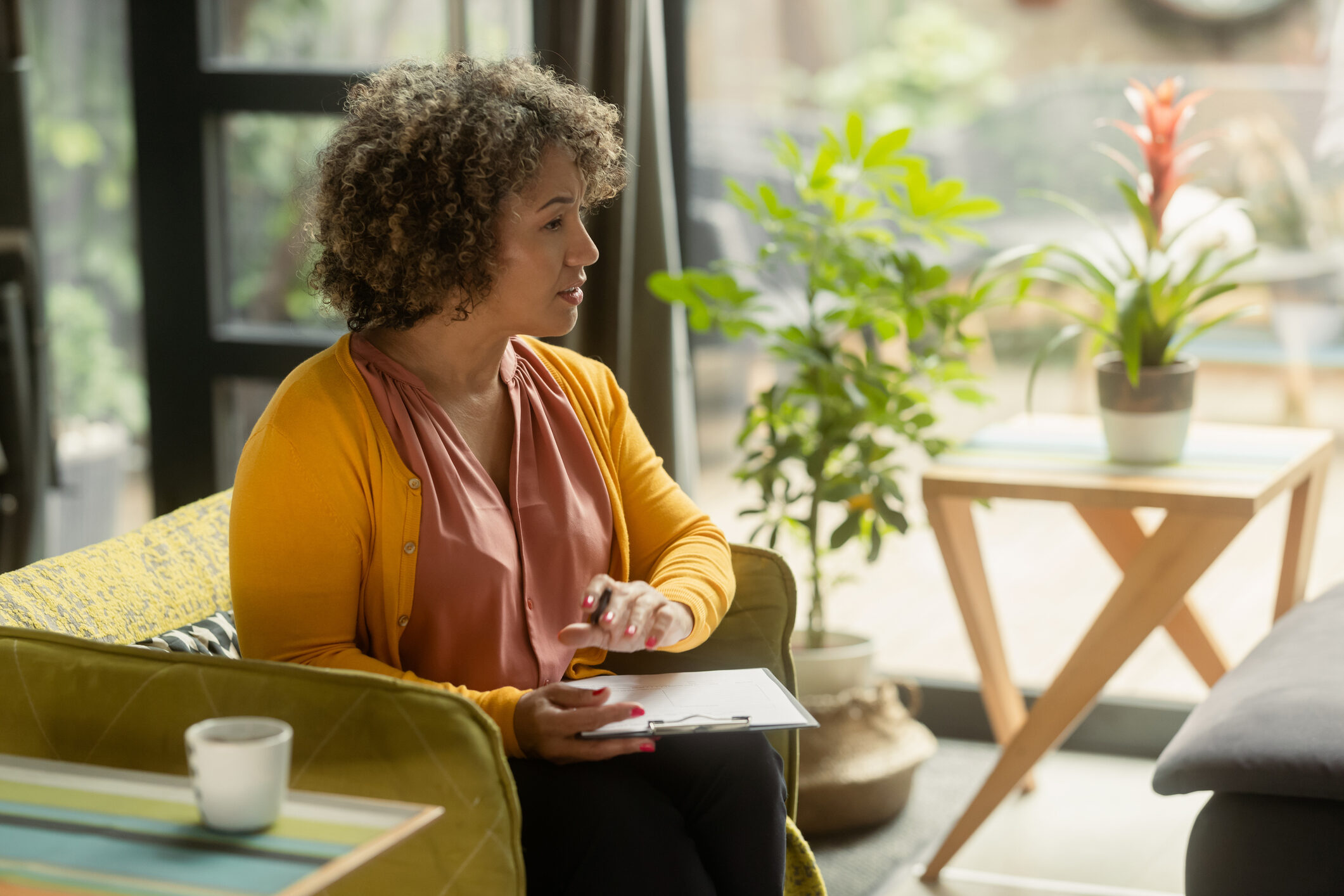 An African-American female counselor sits in the armchair in the office with a clipboard and pen in her hands and listens to the patient.