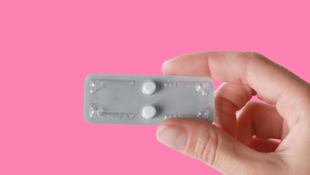 pink background with white complected hand holding two Plan B emergency contraception pills