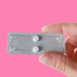 pink background with white complected hand holding two Plan B emergency contraception pills