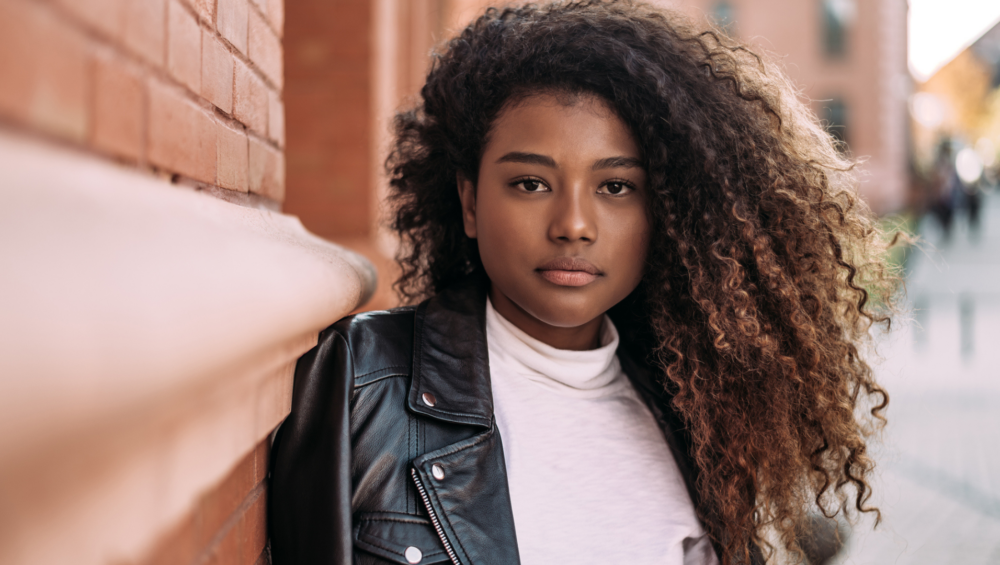 Image of a young beautiful African American woman leaning against a brick wall contemplating how to tell her parents she's pregnant. This image is the header image for the Problem Pregnancy Center.