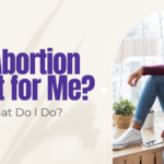 Young woman in a maroon sweater sitting at a window with a phone nearby — thinking about whether abortion is the right choice in Detroit.